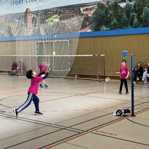Zwei Kinder spielen Badminton in einer Sporthalle, während Zuschauer auf einer Bank im Hintergrund sitzen.