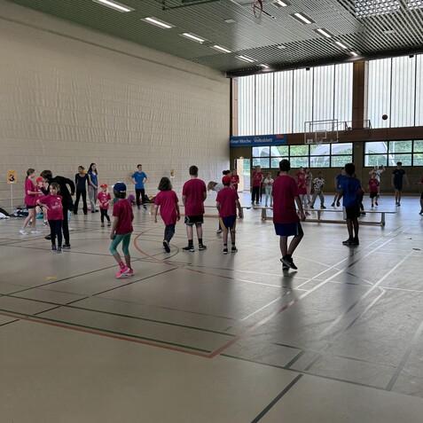 Gruppen von Kindern und Betreuern in roten T-Shirts spielen in einer Sporthalle mit verschiedenen Ballspielen.