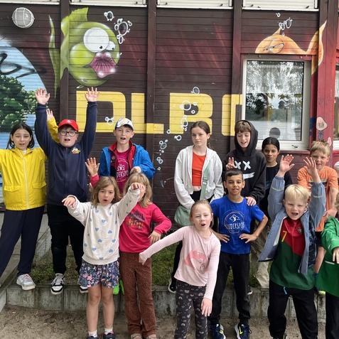 Gruppe von Kindern mit erhobenen H&auml;nden vor einem bunt bemalten Geb&auml;ude mit einer Fisch-Grafik im Hintergrund.