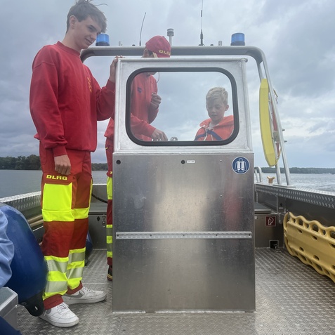 Jugendliche in roten DLRG-Outfits stehen auf einem Rettungsboot, schauen durch ein Fenster und unterhalten sich.