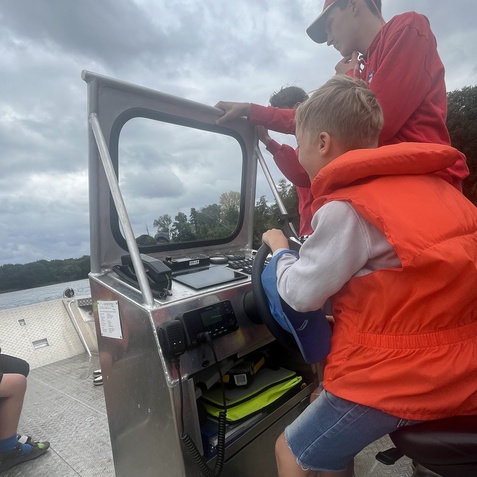 Junge in Rettungsweste lenkt ein Boot, w&auml;hrend ein weiterer Junge ihm &uuml;ber die Schulter schaut. Bew&ouml;lkter Himmel im Hintergrund.