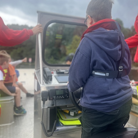 Junge mit Brille steuert ein kleines Boot, w&auml;hrend andere Kinder im Hintergrund sitzen. Wolkiger Himmel ist sichtbar.