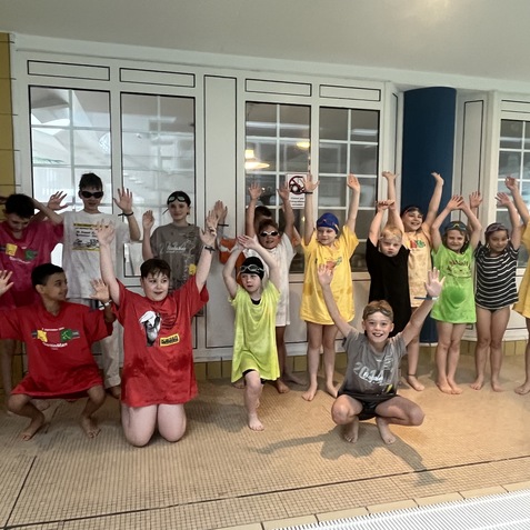Gruppe von Kindern in bunten T-Shirts am Schwimmbad, stehen mit erhobenen H&auml;nden und l&auml;cheln fr&ouml;hlich.
