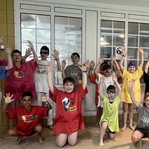 Gruppe von Kindern in bunten T-Shirts und Schwimmbrillen, die fr&ouml;hlich mit erhobenen H&auml;nden posieren.