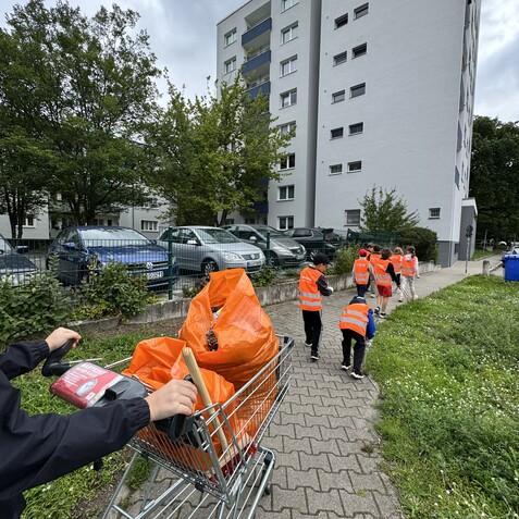Gruppe von Kindern in leuchtend orangefarbenen Westen sammelt M&uuml;ll mit einem Einkaufswagen auf einem Gehweg.
