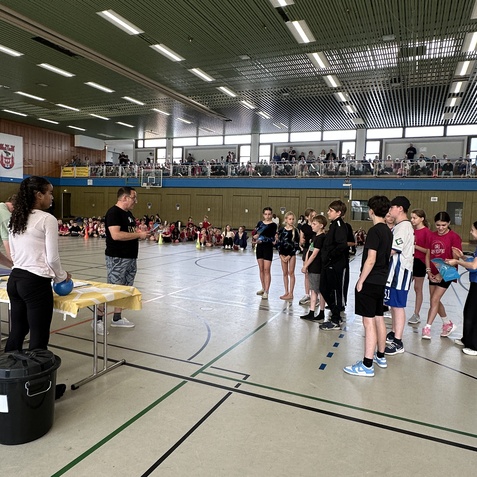 Gruppenszenen in einer Sporthalle mit Jugendlichen beim Warten auf Anweisungen eines Trainers vor einer bunten Publikumskulisse.
