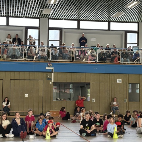 Vielzahl von Zuschauern auf einer Trib&uuml;ne, w&auml;hrend Kinder auf dem Boden einer Turnhalle sitzen und gespannt zuschauen.