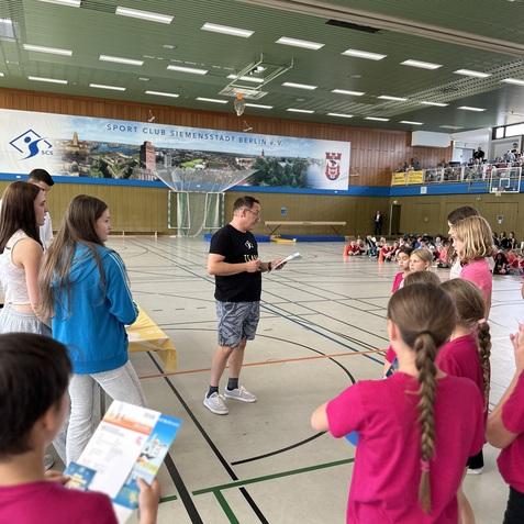 Eine Gruppe von Kindern in pinken T-Shirts h&ouml;rt einem Mann mit Notizen in einer Sporthalle zu.