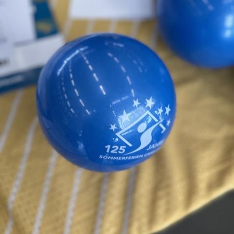 Blauer Ball mit Aufdruck "125 Sommertage" auf gelbem, gestreiften Untergrund. Weitere B&auml;lle im Hintergrund sichtbar.