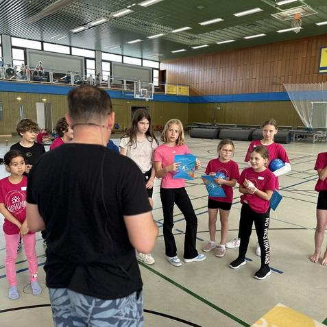 Kinder in pinken T-Shirts stehen in einem Sporthallenraum und h&ouml;ren einem Trainer zu, der Informationen gibt.