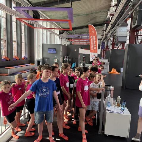 Gruppe von Kindern in bunten T-Shirts wartet in einer Sporthalle, w&auml;hrend eine Betreuerin mit Mikrofon spricht.
