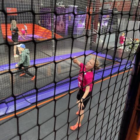 Kind in pinkem Shirt springt auf Trampolin, umgeben von anderen spielenden Kindern und einem Netz.