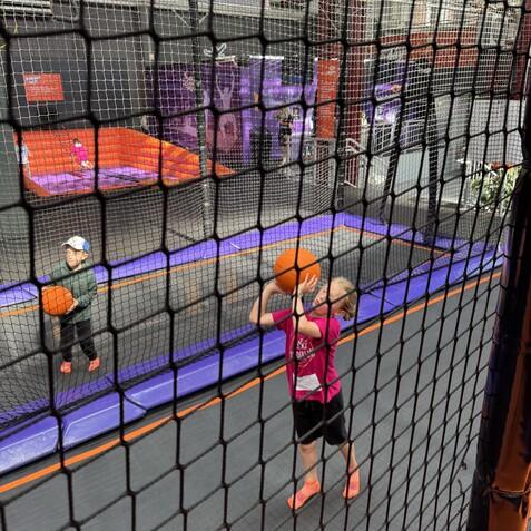 Zwei Kinder spielen mit Basketb&auml;llen auf einem Trampolinplatz, umgeben von einem Sicherheitsnetz.
