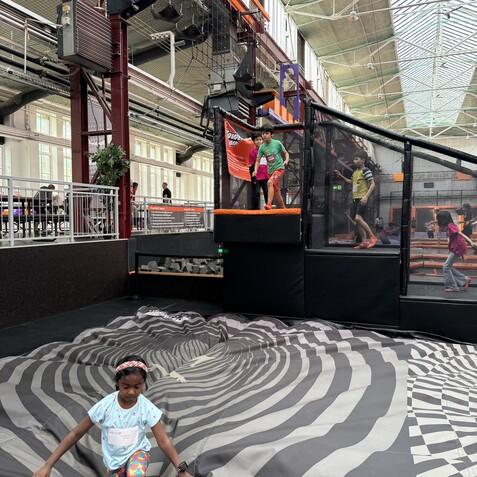 Kind springt auf einer gepolsterten Fl&auml;che mit spiralf&ouml;rmigem Muster in einem Trampolinpark, andere Kinder spielen im Hintergrund.