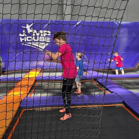 Kinder springen und spielen in einem Trampolinpark, umgeben von einem Netz und bunten Polstern.