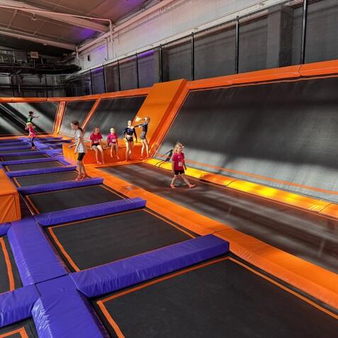Kinder springen und spielen auf Trampolinen in einer bunten Trampolinhalle mit orangefarbenen und lila Elementen.
