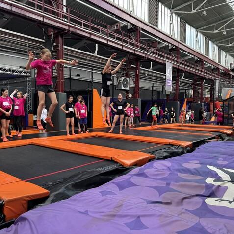 Gruppe von Kindern springt auf Trampolinen in einer Halle mit orangefarbenen und schwarzen Aufprallfl&auml;chen.
