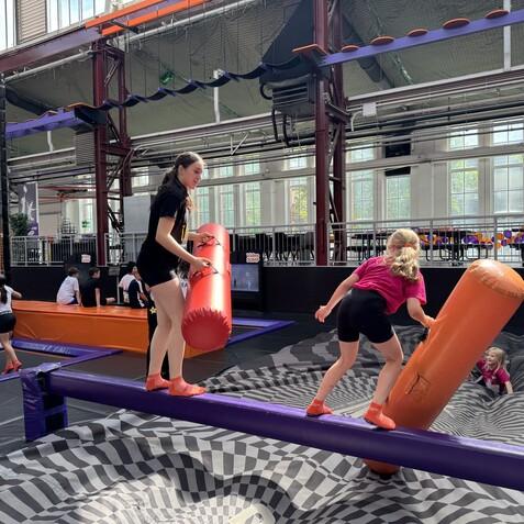 Kinder spielen auf einer Balancebahn in einer Trampolinhalle, w&auml;hrend sie mit gro&szlig;en, gepolsterten Stangen k&auml;mpfen.