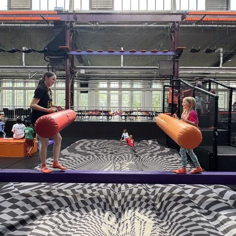 Zwei Kinder mit Schaumstoffstangen balancieren auf einer Plattform in einem Trampolinpark. Im Hintergrund sind weitere Kinder aktiv.