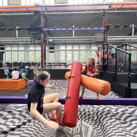 Kinder spielen in einem Trampolinpark, ein M&auml;dchen h&auml;lt eine aufblasbare Stange, w&auml;hrend ein Junge von einer Plattform springt.