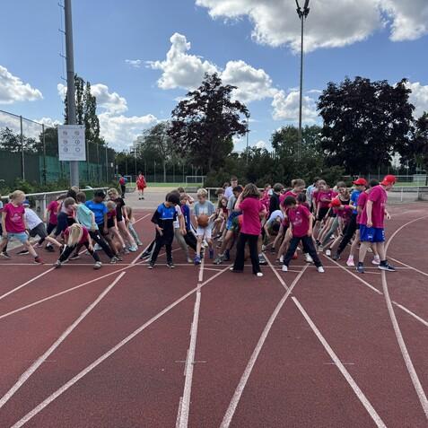 Gruppenspiel von Kindern auf einer Tartanbahn, viele tragen rote T-Shirts und Caps, einige halten einen Basketball.