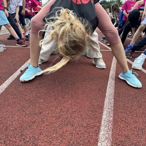 Kinder dehnen sich in Sportkleidung auf einer roten Laufbahn, eine Person in blauen Turnschuhen hebt sich zu einer Dehnung.