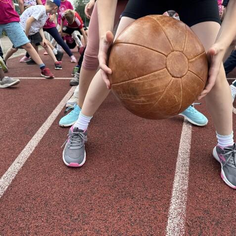 Nahaufnahme von Kindern in Sportkleidung, die auf einer Leichtathletikbahn mit einem schweren Ball trainieren.