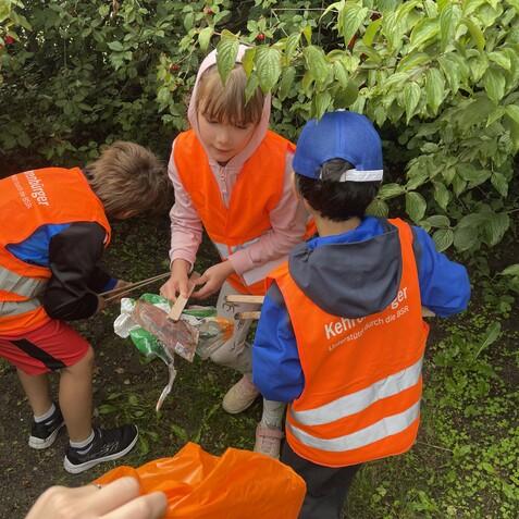 Gruppe von Kindern in orangenen Westen beim Aufr&auml;umen, umgeben von Pflanzen, mit M&uuml;llsammelutensilien in den H&auml;nden.