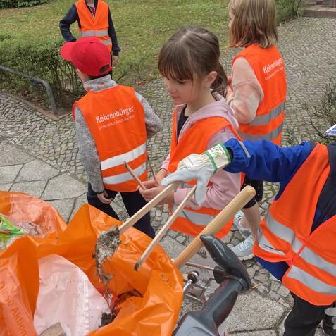 Gruppe von Kindern mit orangefarbenen Westen, die M&uuml;ll in einen orangefarbenen Sack sammeln.
