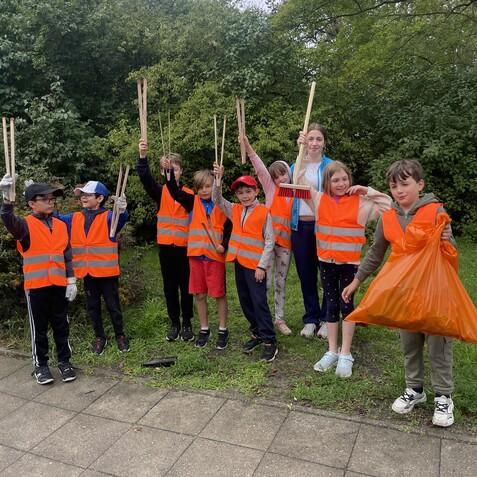 Gruppe von Kindern in orangefarbenen Warnwesten mit M&uuml;llzangen und einem M&uuml;llsack, stehend in einer Gr&uuml;nanlage.