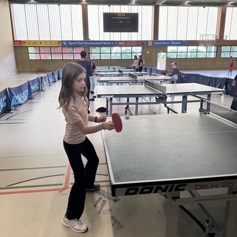 M&auml;dchen mit Tischtennisschl&auml;ger am Tisch, w&auml;hrend andere Spieler im Hintergrund aktiv sind. Sporthalle mit Tischtennistischen.