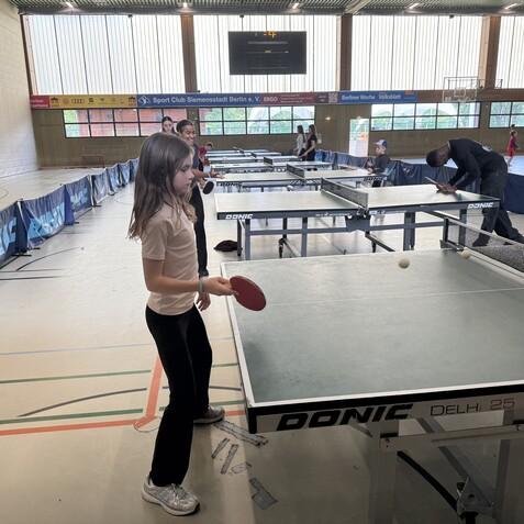 M&auml;dchen mit Tischtennisschl&auml;ger an einem Tisch, w&auml;hrend B&auml;lle &uuml;ber das Netz fliegen in einer Sporthalle.