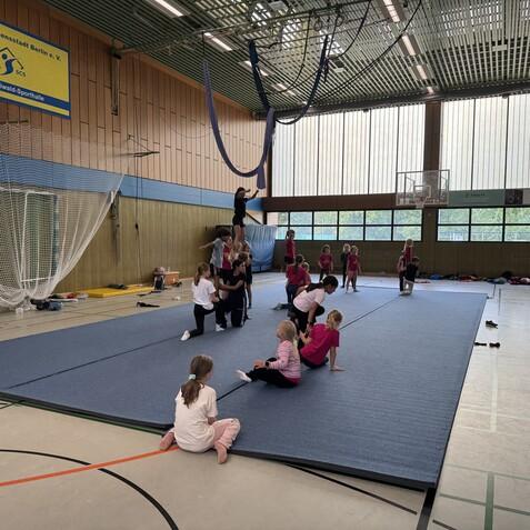 Gruppe von Kindern und Jugendlichen trainiert akrobatische &Uuml;bungen auf einer blauen Gymnastikmatte in einer Sporthalle.