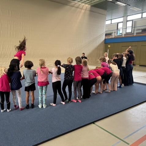 Gruppentraining von Kindern in einer Turnhalle; einige springen, andere stehen in einer Linie auf einem blauen Turnmattenboden.