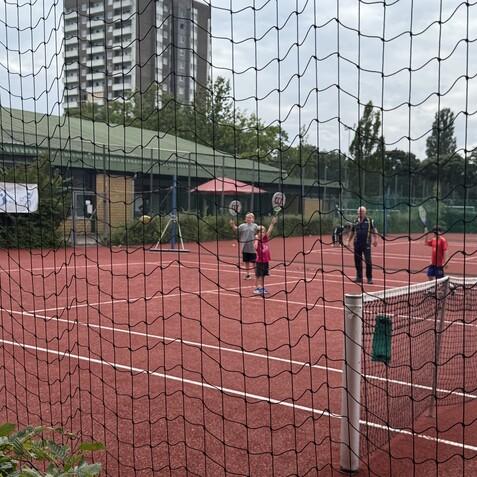 Jugendliche und Erwachsene spielen Tennis auf einem roten Platz, umgeben von einem Netz und gr&uuml;ner Vegetation.