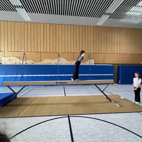 Ein M&auml;dchen springt auf einem Schwebebalken, w&auml;hrend zwei andere M&auml;dchen daneben stehen und zuschauen.