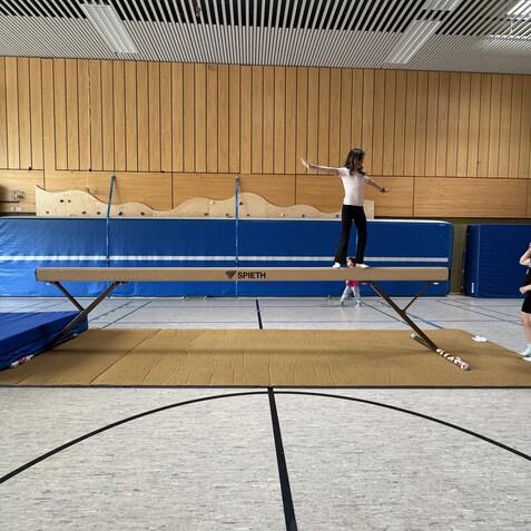 M&auml;dchen balanciert auf einem Schwebebalken in einer Turnhalle, w&auml;hrend zwei weitere M&auml;dchen zuschauen.