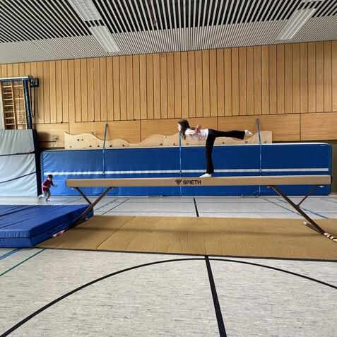 M&auml;dchen balanciert auf einem Schwebebalken in einer Turnhalle, w&auml;hrend zwei andere Kinder im Hintergrund spielen.