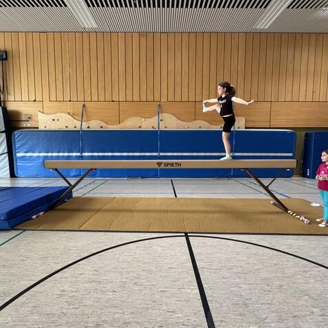 M&auml;dchen balanciert auf einem Schwebebalken in einer Turnhalle, w&auml;hrend zwei andere M&auml;dchen zuschauen.