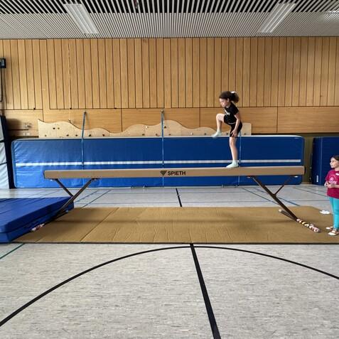 M&auml;dchen turnt auf einem Schwebebalken, w&auml;hrend zwei andere M&auml;dchen und eine Trainerin zuschauen. Sicherheitsmatte liegt darunter.