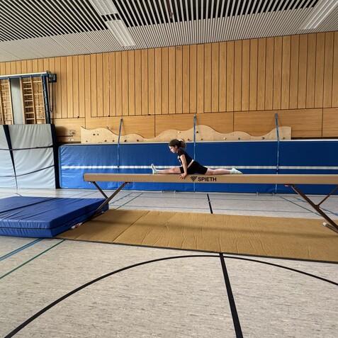 M&auml;dchen beim Turnen auf dem Schwebebalken, ein weiteres M&auml;dchen beobachtet daneben in einer Turnhalle.