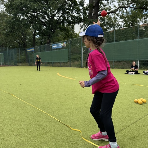 M&auml;dchen in pinkem T-Shirt wirft einen roten Ball auf einem gr&uuml;nen Spielfeld, im Hintergrund Zuschauer und B&auml;ume.