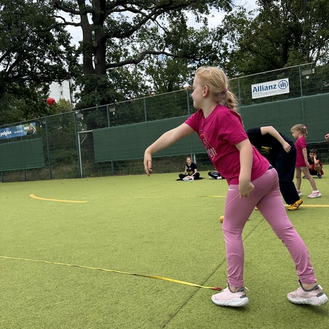 M&auml;dchen in rosa Shirt wirft einen Ball auf einem gr&uuml;nen Sportplatz, andere Kinder sitzen im Hintergrund.