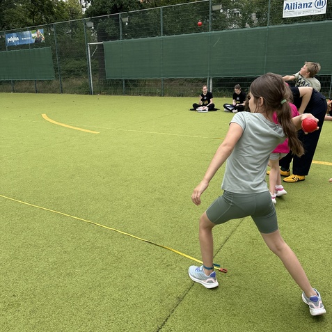 Ein M&auml;dchen wirft einen roten Ball auf einem gr&uuml;nen Sportplatz, w&auml;hrend andere Kinder zuschauen.