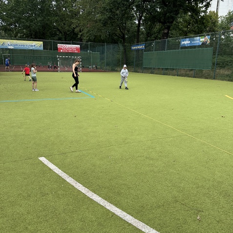 Zwei Kinder und eine Erwachsene auf einem Kunstrasenplatz, umgeben von B&auml;umen und Werbebannern im Hintergrund.