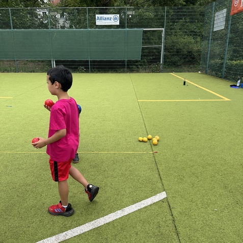 Zwei Kinder stehen auf einem Kunstrasenplatz, eines h&auml;lt einen roten Ball, w&auml;hrend gelbe B&auml;lle am Boden liegen.