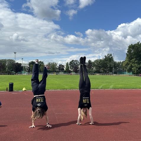 Zwei Personen machen Handstand auf einem roten Sportplatz, mit gr&uuml;nem Rasen und wolkigem Himmel im Hintergrund.