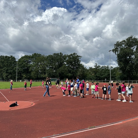 Gruppe von Kindern in Sportkleidung &uuml;bt auf einem roten Sportplatz unter bew&ouml;lktem Himmel.