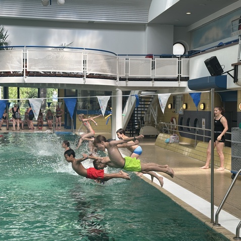 F&uuml;nf Kinder springen ins Wasser eines Schwimmbeckens, w&auml;hrend eine Trainerin am Beckenrand zusieht.