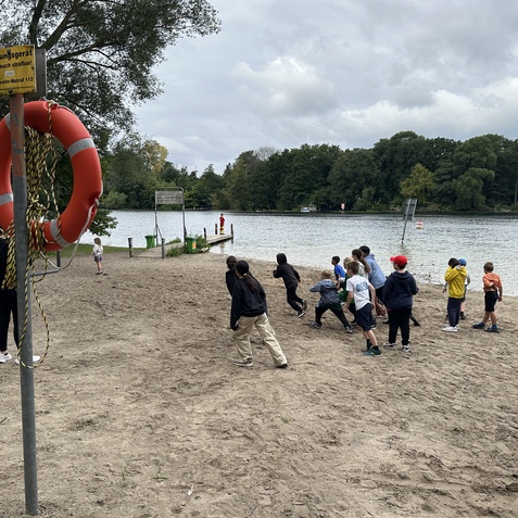 Gruppe von Kindern spielt am Sandstrand eines Gew&auml;ssers, mit einem Rettungsring und einem Steg im Hintergrund.